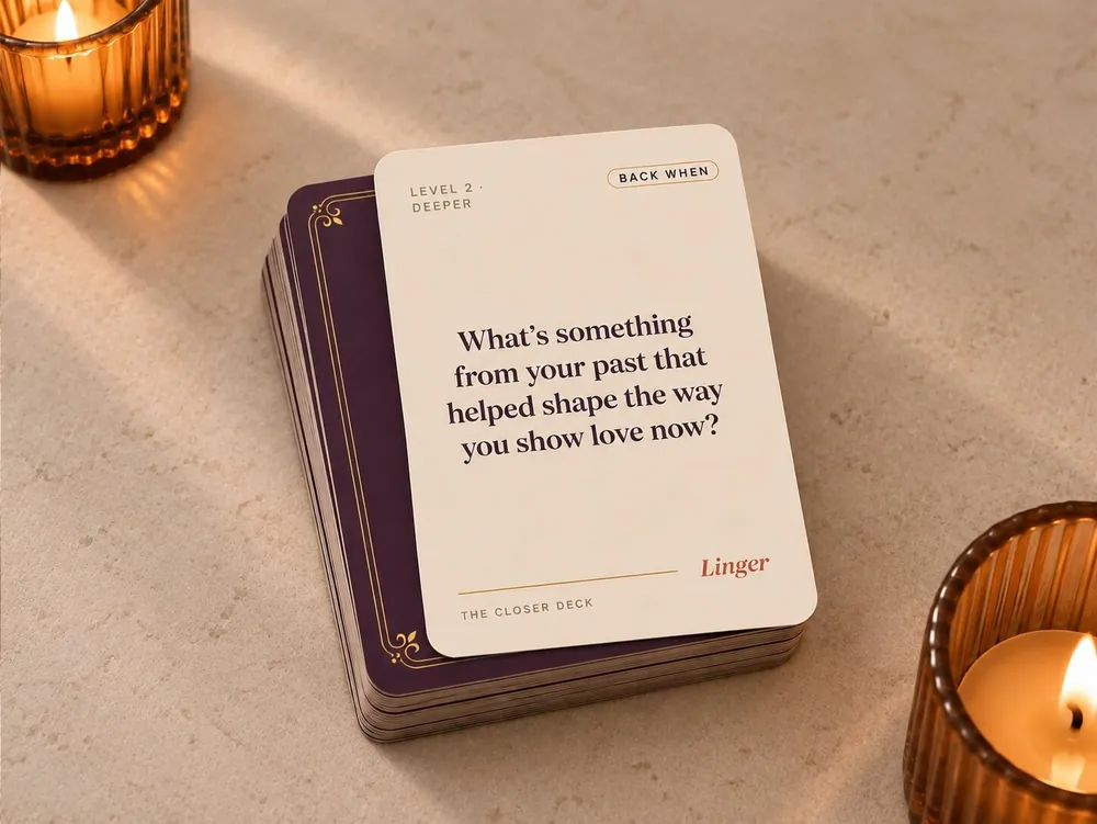 The Linger Closer Deck conversation cards for couples — a cream question card reading 'What's something from your past that helped shape the way you show love now?' resting on a stack of plum cards between two candles.